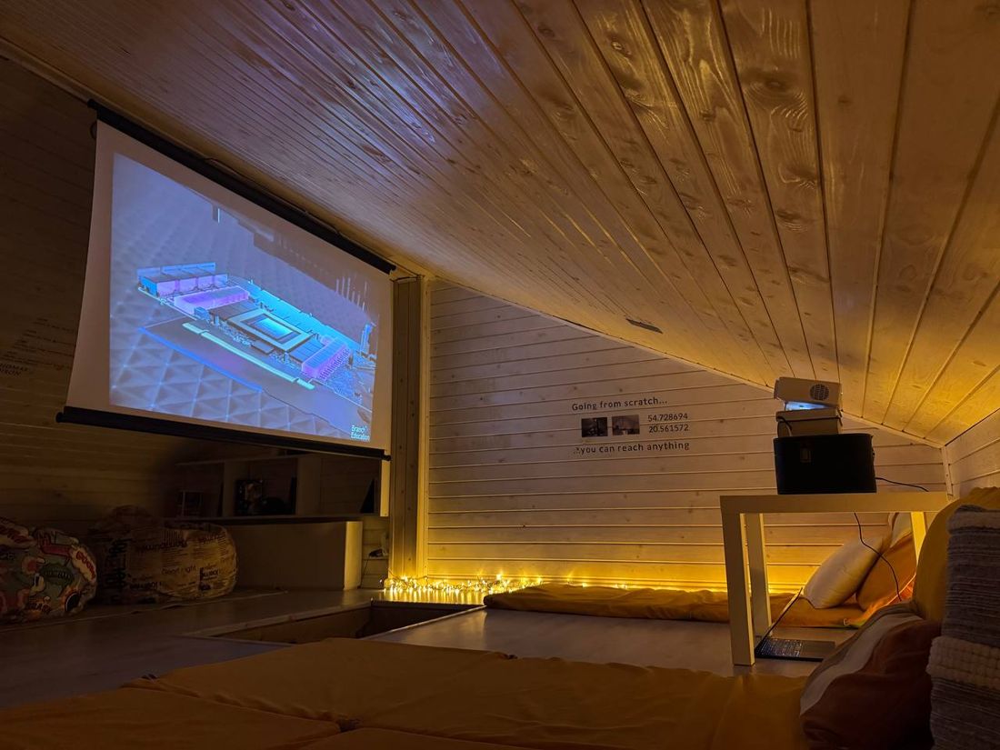 The lounge in warm light: mats, string lights, a projector and a screen showing a chip being built